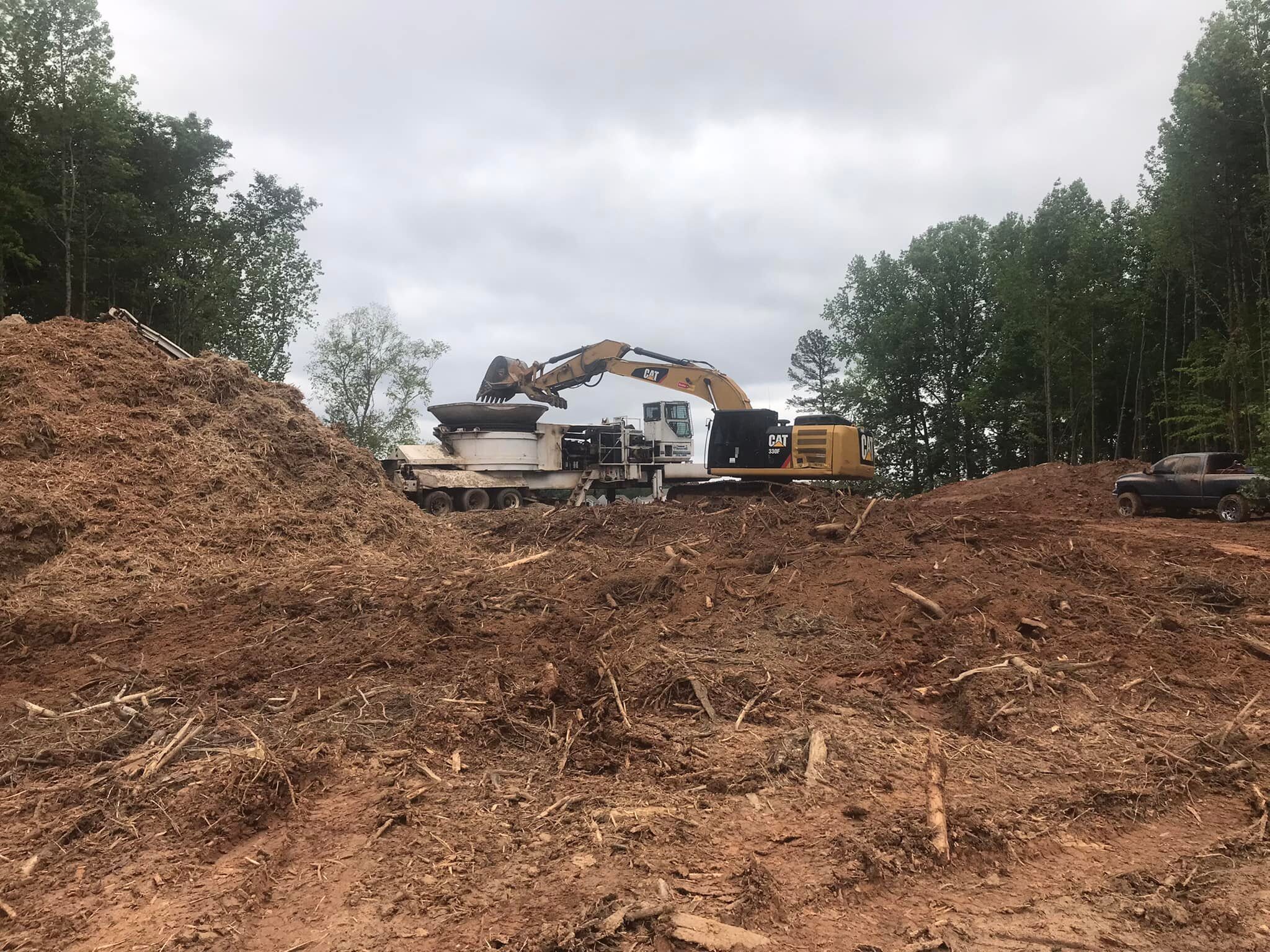 The Elder's Grinder in action, on top of a hill of mulch with a cloudy, overcast sky in the background, and flanked by tree lines on both sides.